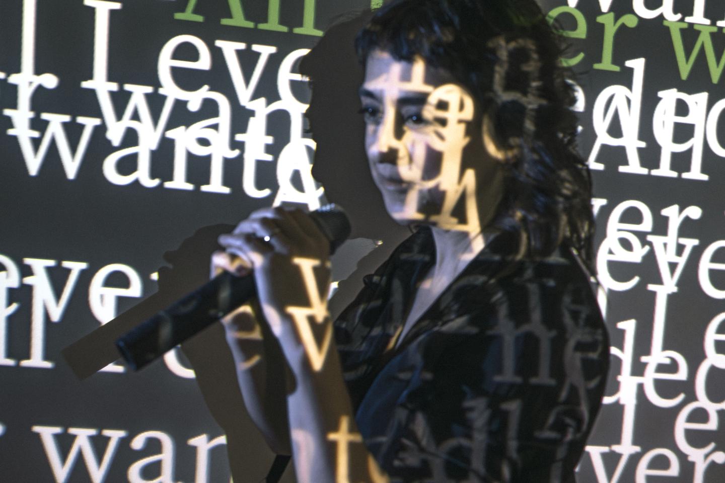 Zdjęcie kobiety trzymającej mikrofon. Na jej postaci i dookoła niej wyświetlane są napisy z projektora.