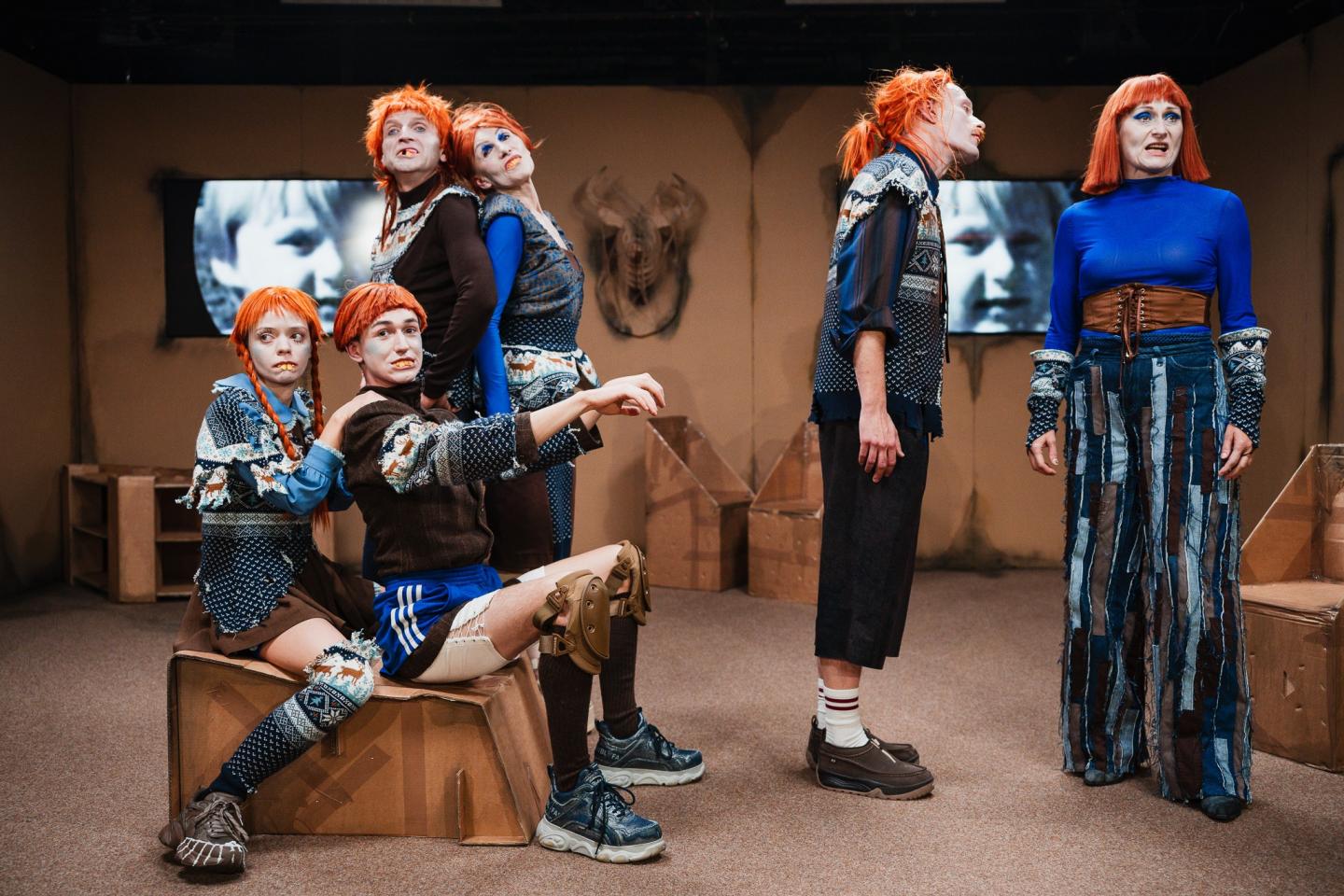 Grupa aktorów w kartonowej scenografii. Na ścianie dwa telewizory, wyświetlające stare zdjęcie małej dziewczynki. Aktorzy mają na sobie rude peruki i kostiumy z niebieskimi elementami. Z przodu na kartonowym siedzisku siedzą młoda kobieta i młody mężczyzna. 