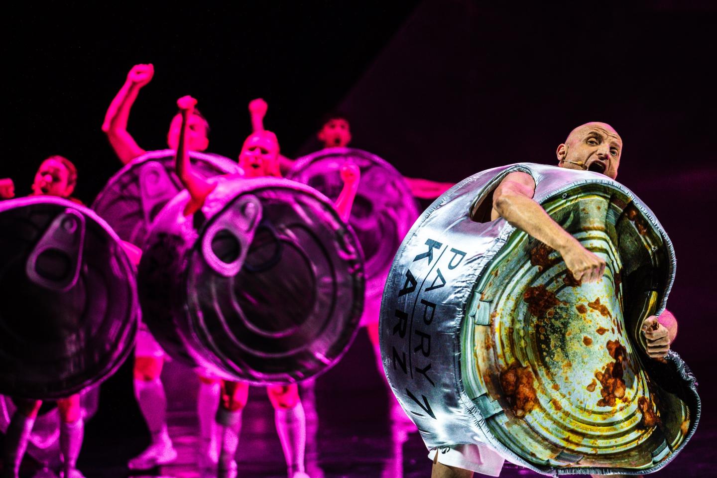 Mężczyzna w stroju wielkiej aluminiowej puszki. Puszka wygląda jakby była otwarta i pusta, zabrudzona resztkami jedzenia. Za nim grupa osób unoszących dłonie ku górze, podświetlona różowym światłem.