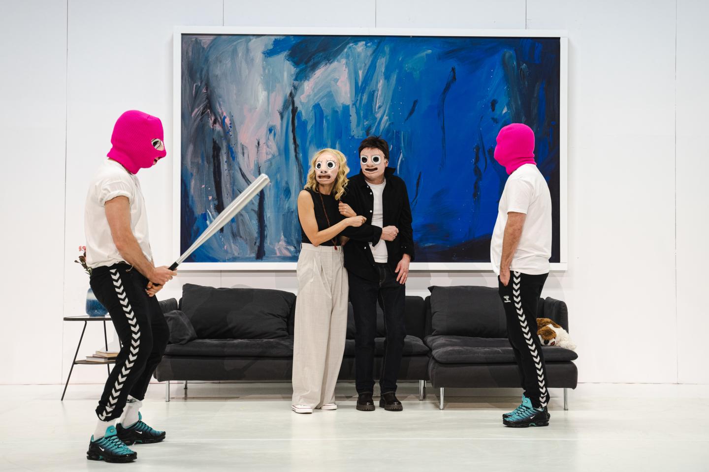 Dwójka mężczyzn ubranych w różowe kominiarki, białe bluzki i czarne spodnie dresowe, stoi po obu stronach zdjęcia. Między nimi stoi przestraszona para- mężczyzna i kobieta. W tle jest szara kanapa i niebieski obraz. 