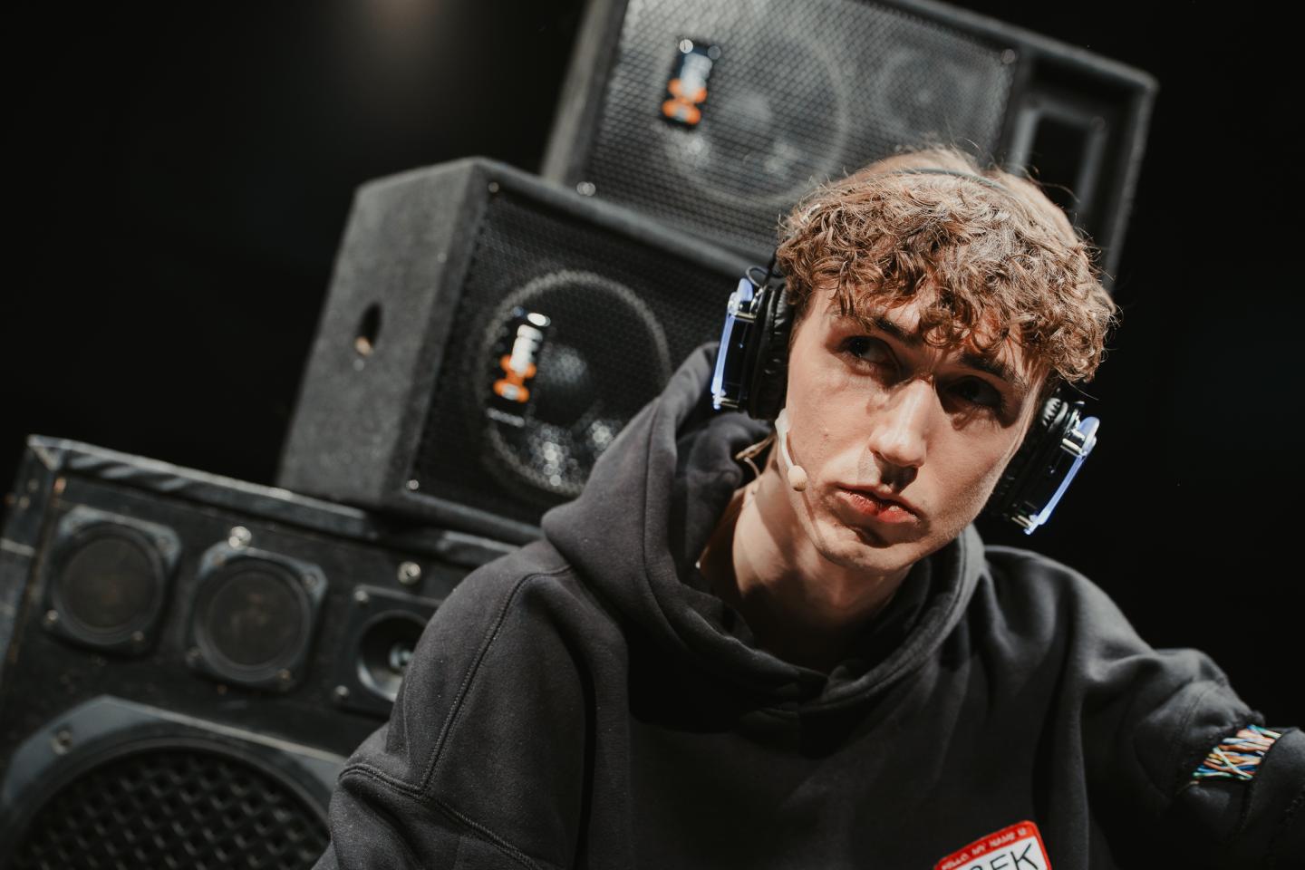 Młody mężczyzna w dużych słuchawkach nausznych. Ubrany w czarną bluzę z kapturem. Spogląda w dal. W tle widać duże czarne głośniki ustawione jeden na drugim.