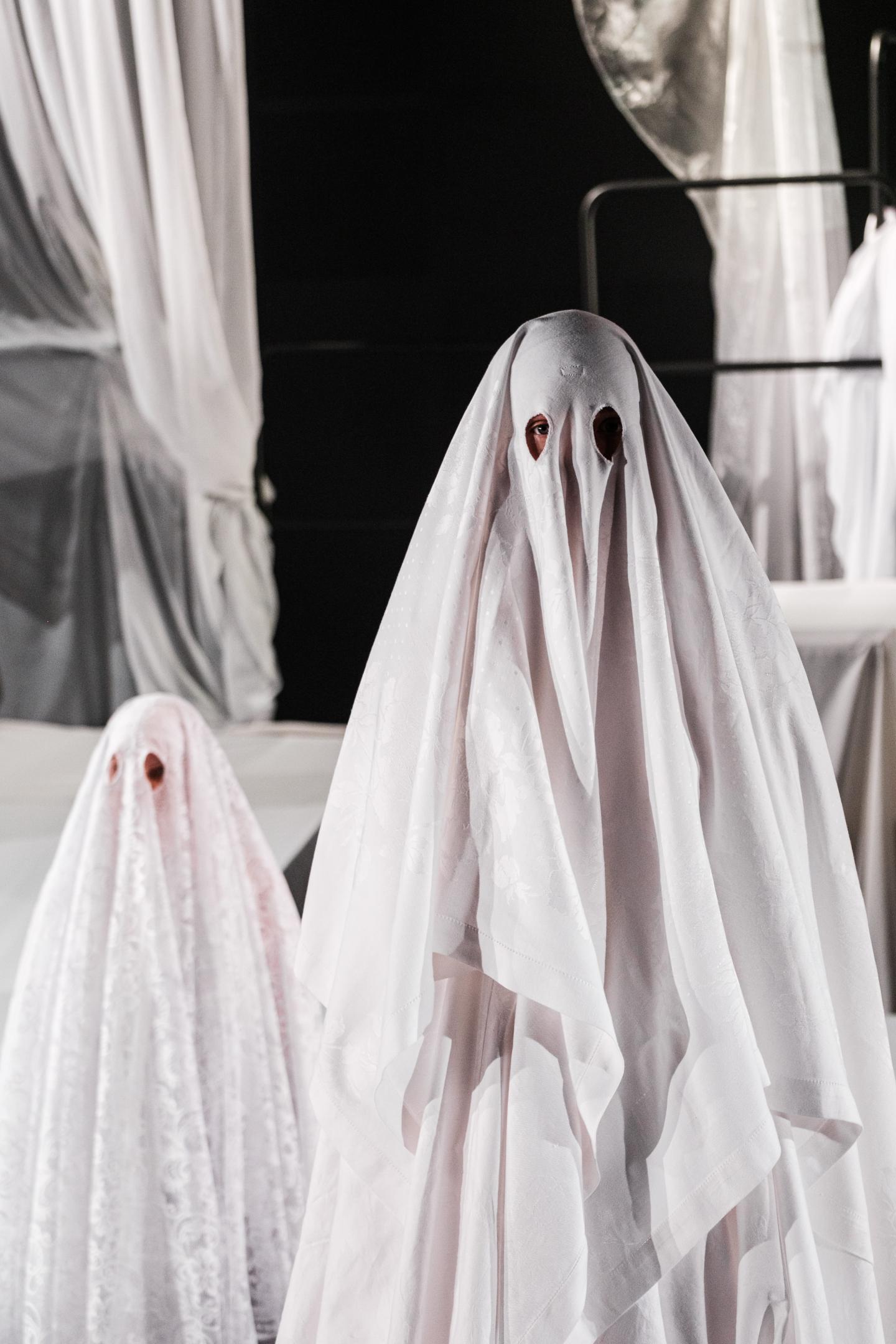 Dwie postacie ubrane w prześcieradła przedstawiające duchy. W tle widać białe zasłony na czarnym tle. 