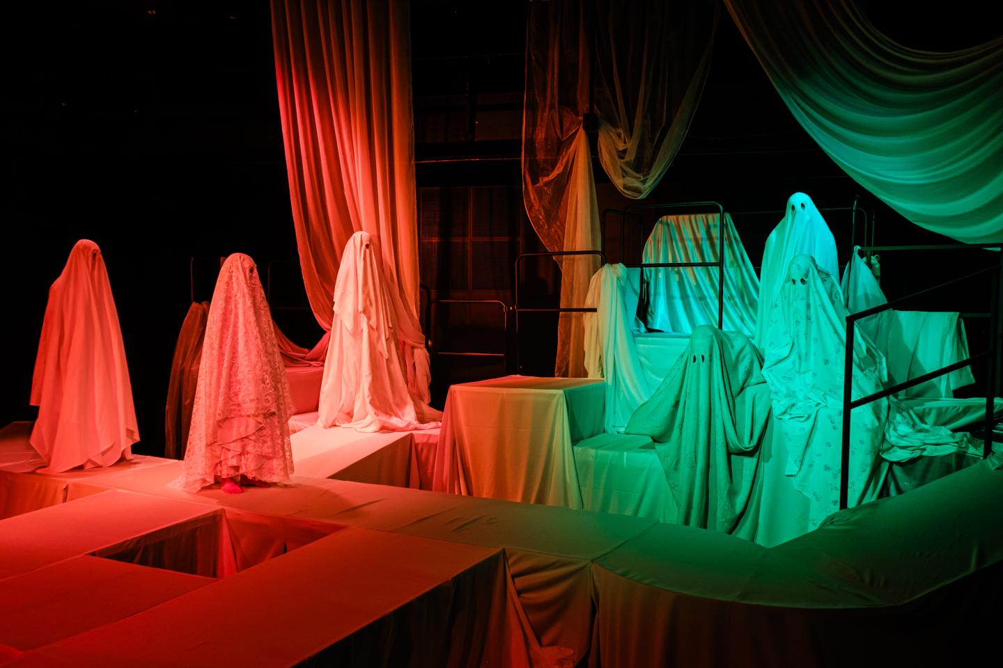 Zdjęcie przedstawia grupę postaci w białych prześcieradłach przebranych za duchy. Stoją na platformach. Zdjęcie jest oświetlone na czerwono i zielono. 