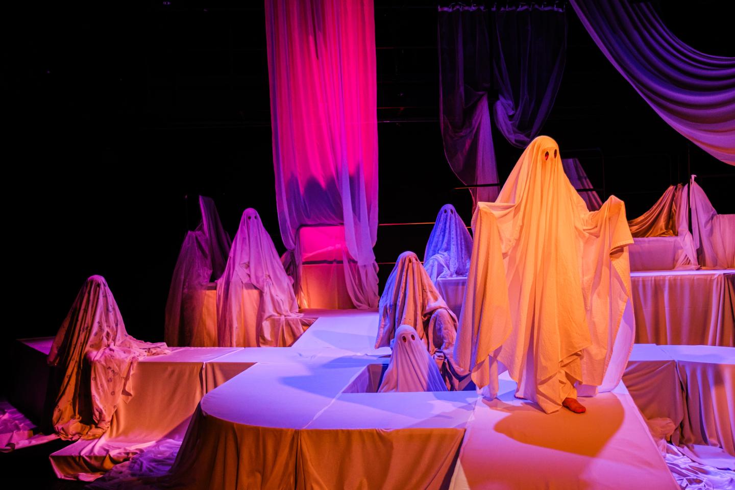 Zdjęcie przedstawia grupę postaci przebranych w prześcieradła jako duchy. Scena jest oświetlona na różowo i pomarańczowo. 